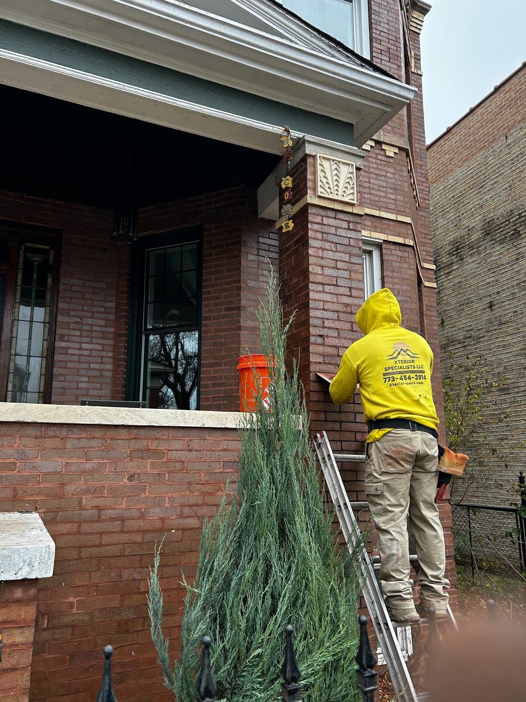 Crew member in a yellow Xterior Specialists hoodie on a ladder tuckpointing a brick porch column.
