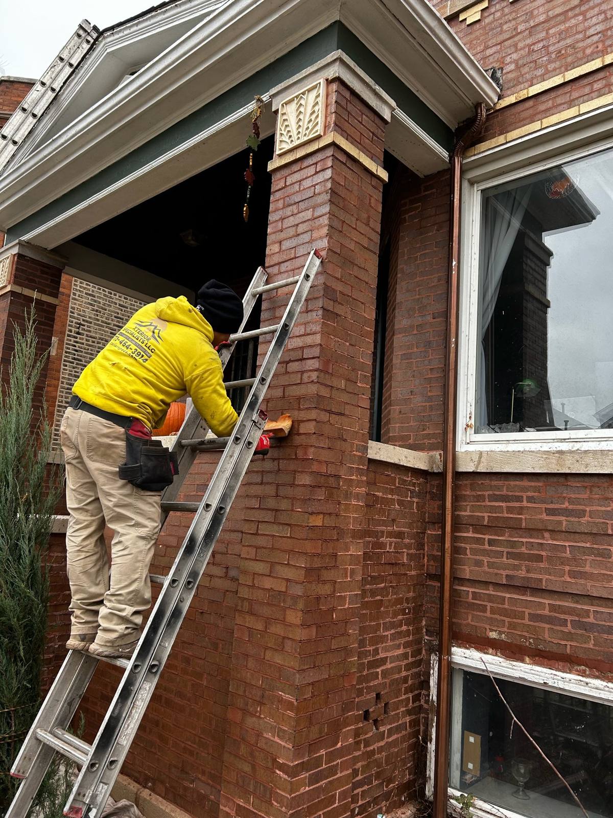 Crew member in a yellow Xterior Specialists hoodie on an extension ladder working the mortar joints of a brick porch column.
