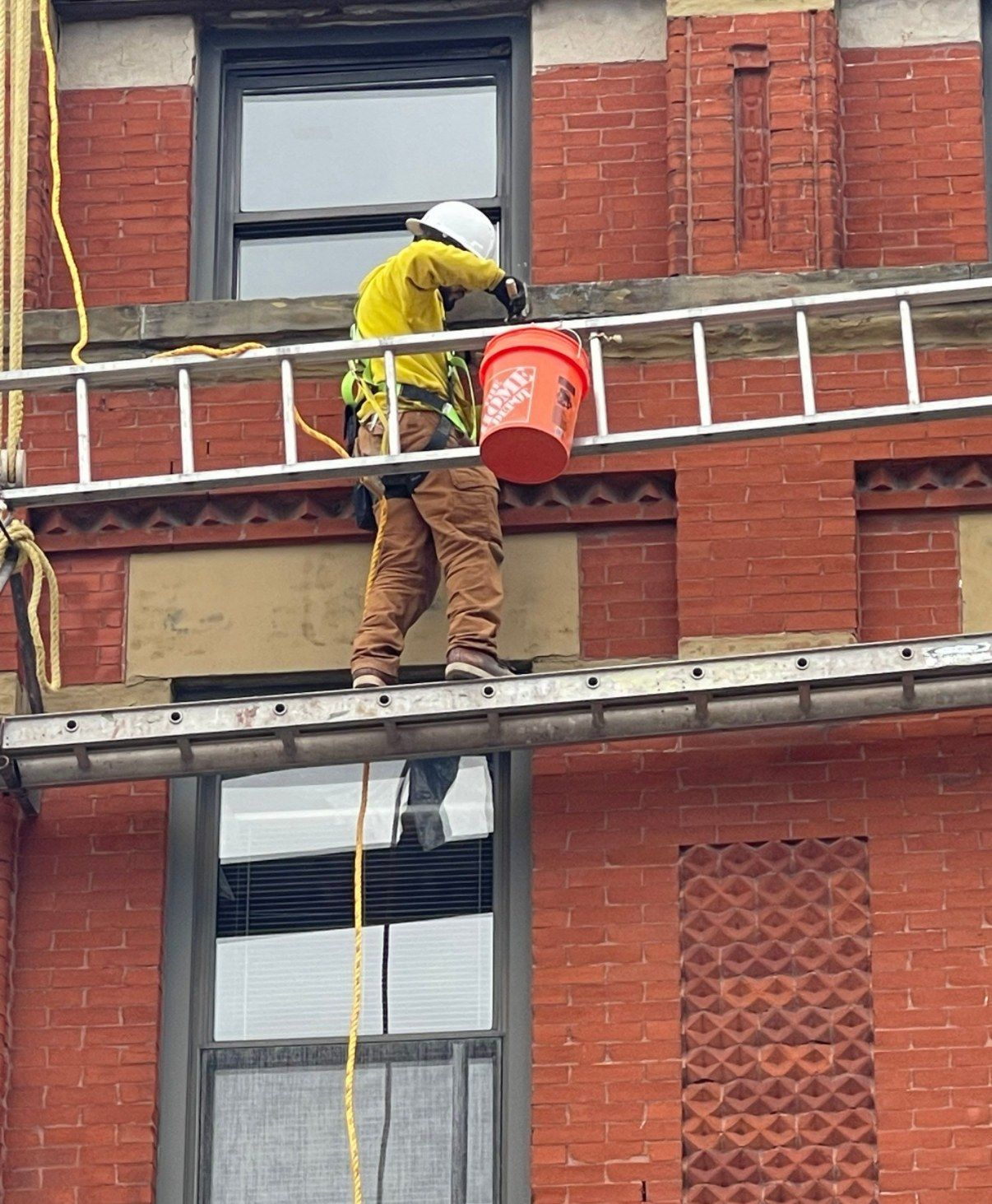 Crew member in a hard hat and harness working from a ladder scaffold on the face of a multi-story red brick building, between window openings.