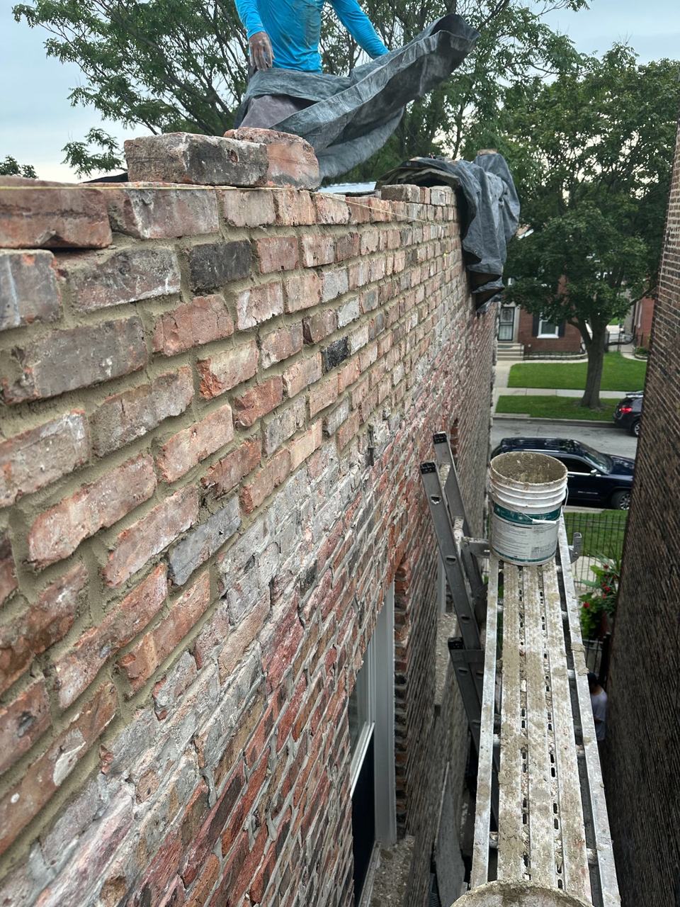 Brick parapet being rebuilt with fresh mortar joints, with a plank scaffold and ladder set up along the side of the building.