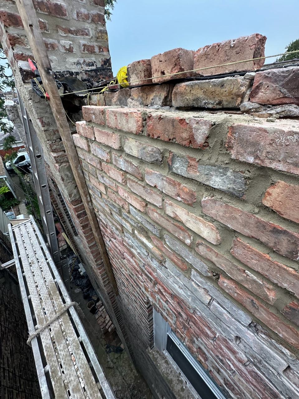Freshly laid brick courses at the top of a parapet being rebuilt, with a string line and scaffold planks alongside.