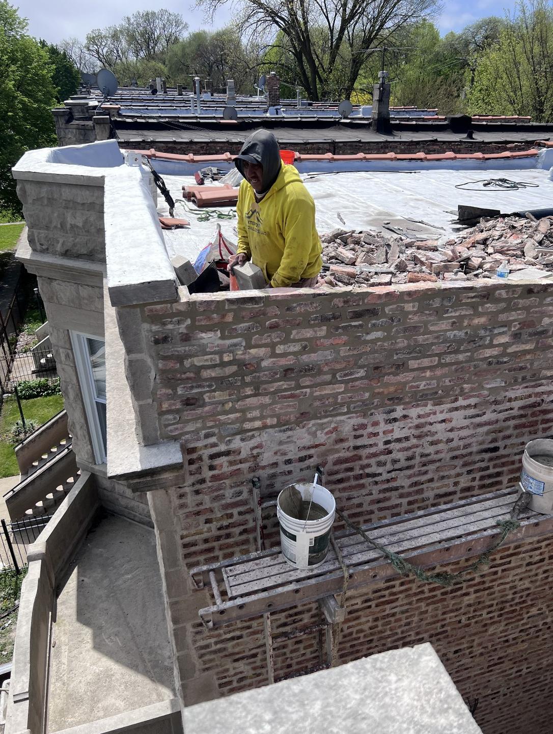 Crew member in a yellow Xterior Specialists hoodie rebuilding a brick parapet on a flat roof, with old brick piled on the roof and a plank scaffold along the wall.