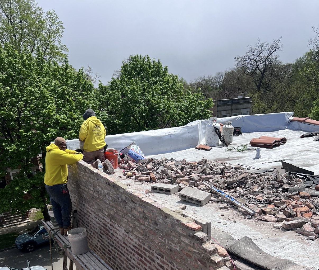 Two crew members in yellow hoodies working on a brick parapet from a plank scaffold, with old brick removed from the wall piled on the flat roof.