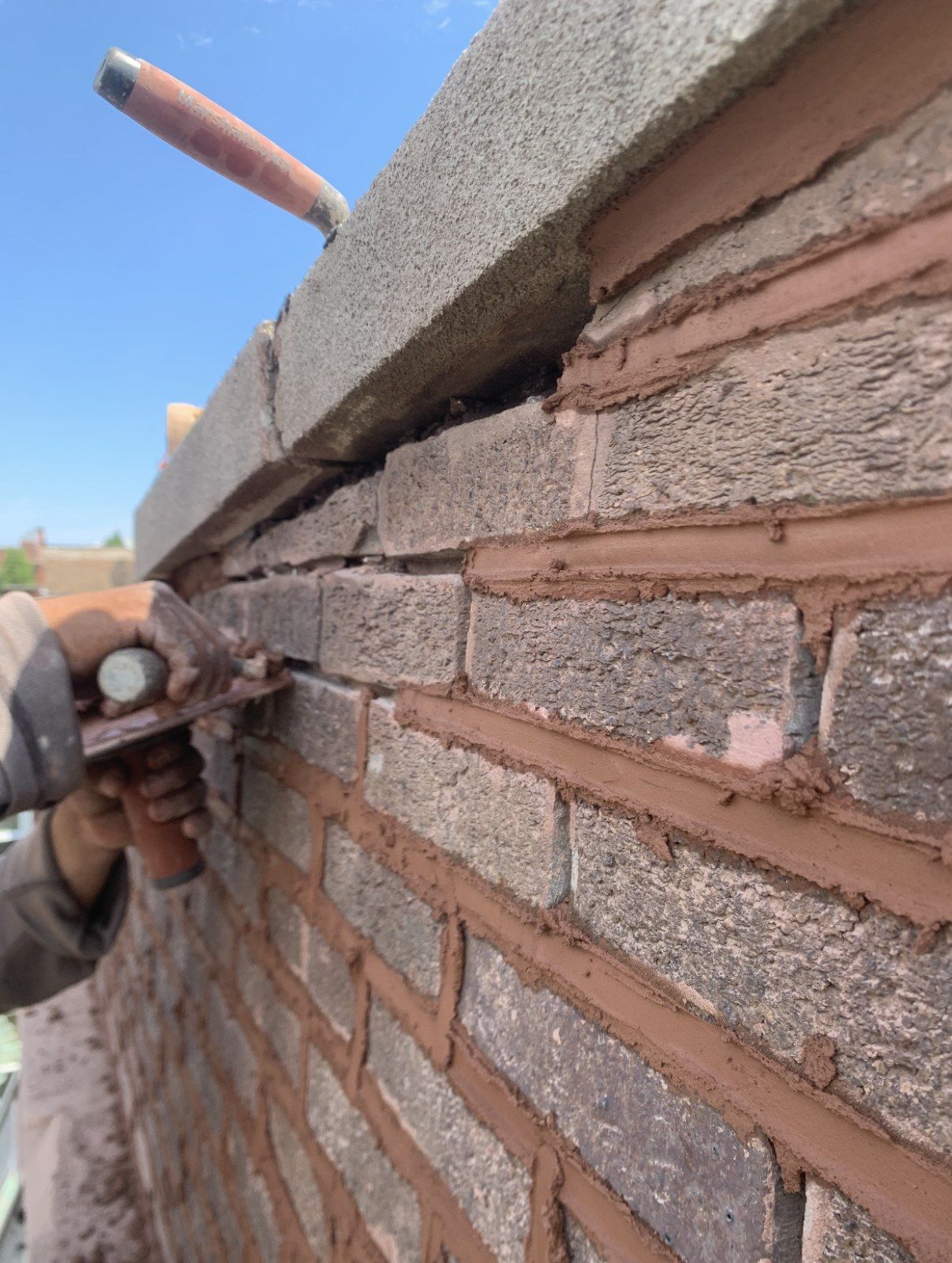 Gloved hand tooling fresh red mortar into brick joints directly below stone coping, with a trowel resting on top.