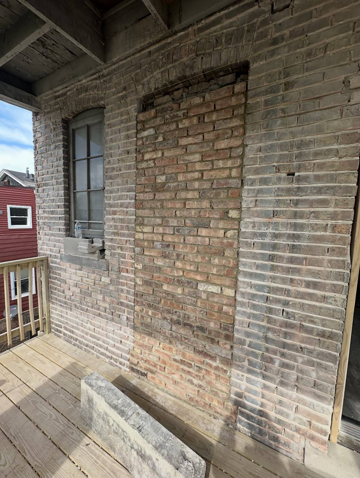 Former window opening on a brick wall filled in with brick beneath the original brick arch, next to an existing window on a rear porch.